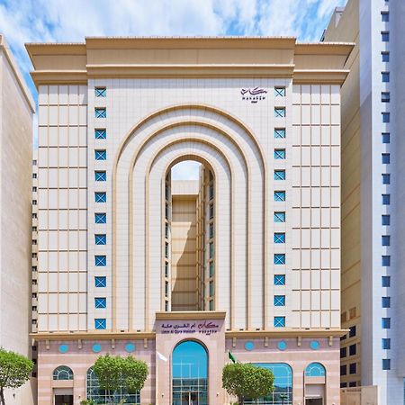 Makarem Umm Al Qura Hotel Mecca Exterior foto The photo shows a tall, modern building with a distinctive architectural design. The facade features a series of large arches and has a light-colored exterior with decorative patterns. There are blue accents that provide contrast against the neutral 