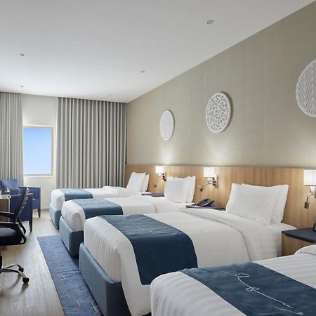 Makarem Umm Al Qura Hotel Mecca Exterior foto The photo shows a modern hotel room featuring three beds, all neatly made with white linens and blue accents. The room has soft, neutral-colored walls and wooden elements. There is a large television mounted on the wall opposite the beds and a work d