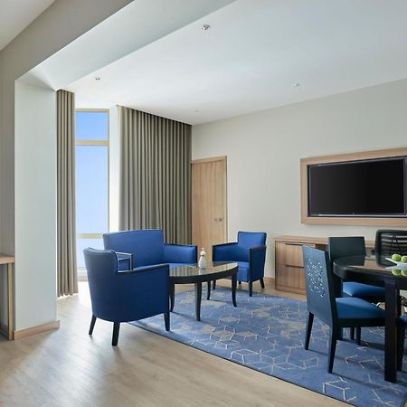 Makarem Umm Al Qura Hotel Mecca Exterior foto The photo shows a spacious and modern living area. It features a set of blue armchairs arranged around a glass coffee table on a patterned rug. There is a large window with flowing curtains that lets in natural light, giving the space a bright and ai