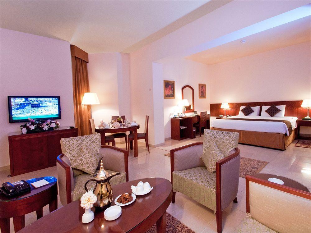 Makarem Umm Al Qura Hotel Mecca Exterior foto The photo shows a spacious hotel room featuring a well-appointed interior. In the foreground, there are two comfortable chairs and a small table with a decorative centerpiece and snacks. To the left, a television is mounted on the wall, providing ent