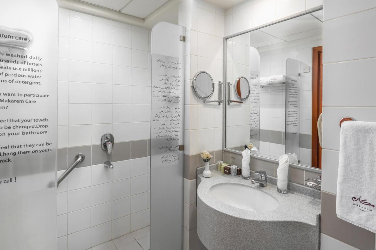 Makarem Umm Al Qura Hotel Mecca Exterior foto The photo shows a modern bathroom with a clean, minimalist design. It features a shower area separated by a glass panel, and there is a round mirror above a curved sink. The sink is accompanied by various toiletries and a hand towel. White tiles domi