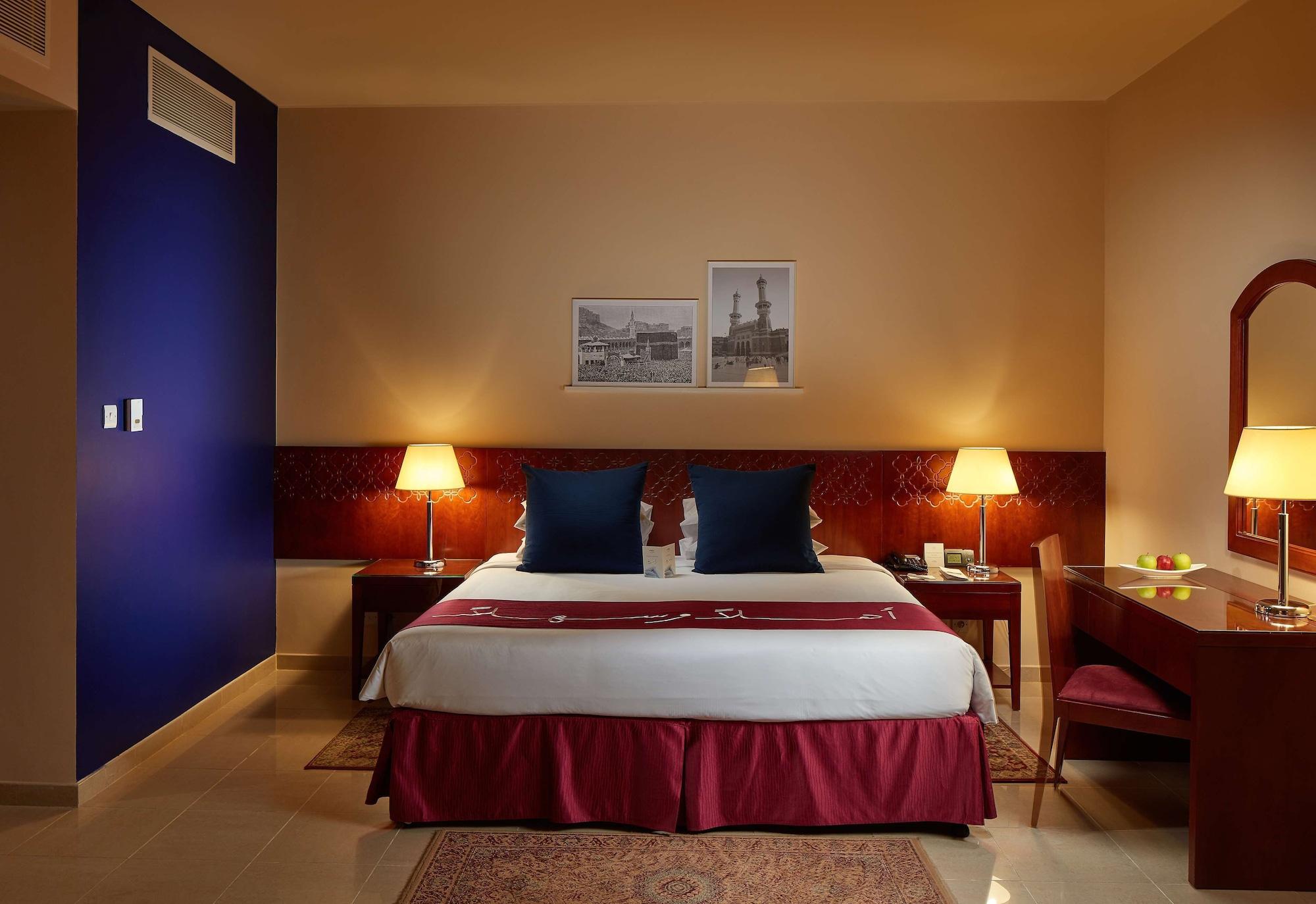 Makarem Umm Al Qura Hotel Mecca Exterior foto The photo shows a hotel room with a modern and cozy design. It features a large bed with a white duvet and burgundy bed skirt, complemented by two dark blue pillows. On either side of the bed, there are lamps providing soft lighting. The walls are pa