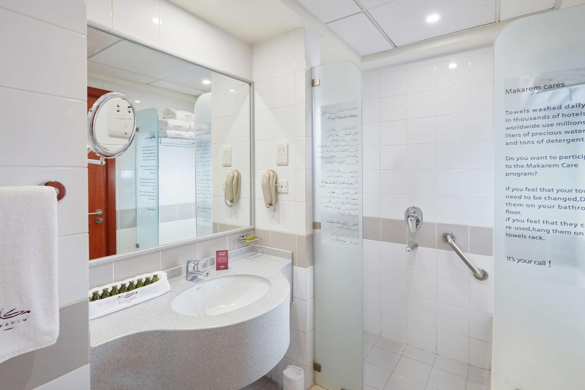 Makarem Umm Al Qura Hotel Mecca Exterior foto The photo shows a modern bathroom with a clean and minimalist design. There is a curved vanity sink with a large mirror above it, which also includes a circular mirror attached to the wall. The sink area has a few toiletries displayed. On one side, t