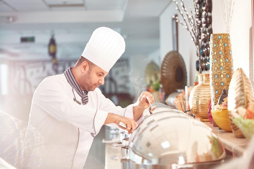 Makarem Umm Al Qura Hotel Mecca Exterior foto The photo shows a chef in a white uniform and tall chef's hat, focused on preparing food at a buffet or catering station. He is leaning over steaming food trays, indicating that he is either serving or checking the dishes. The background features dec