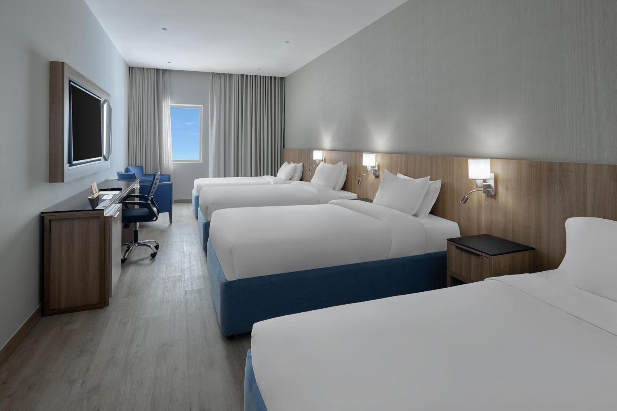 Makarem Umm Al Qura Hotel Mecca Exterior foto The photo shows a modern hotel room featuring four beds set up in a spacious layout. Each bed has white linens and blue accents. There is a large flat-screen TV mounted on the wall, and a desk with a chair is positioned next to it, providing a worksp