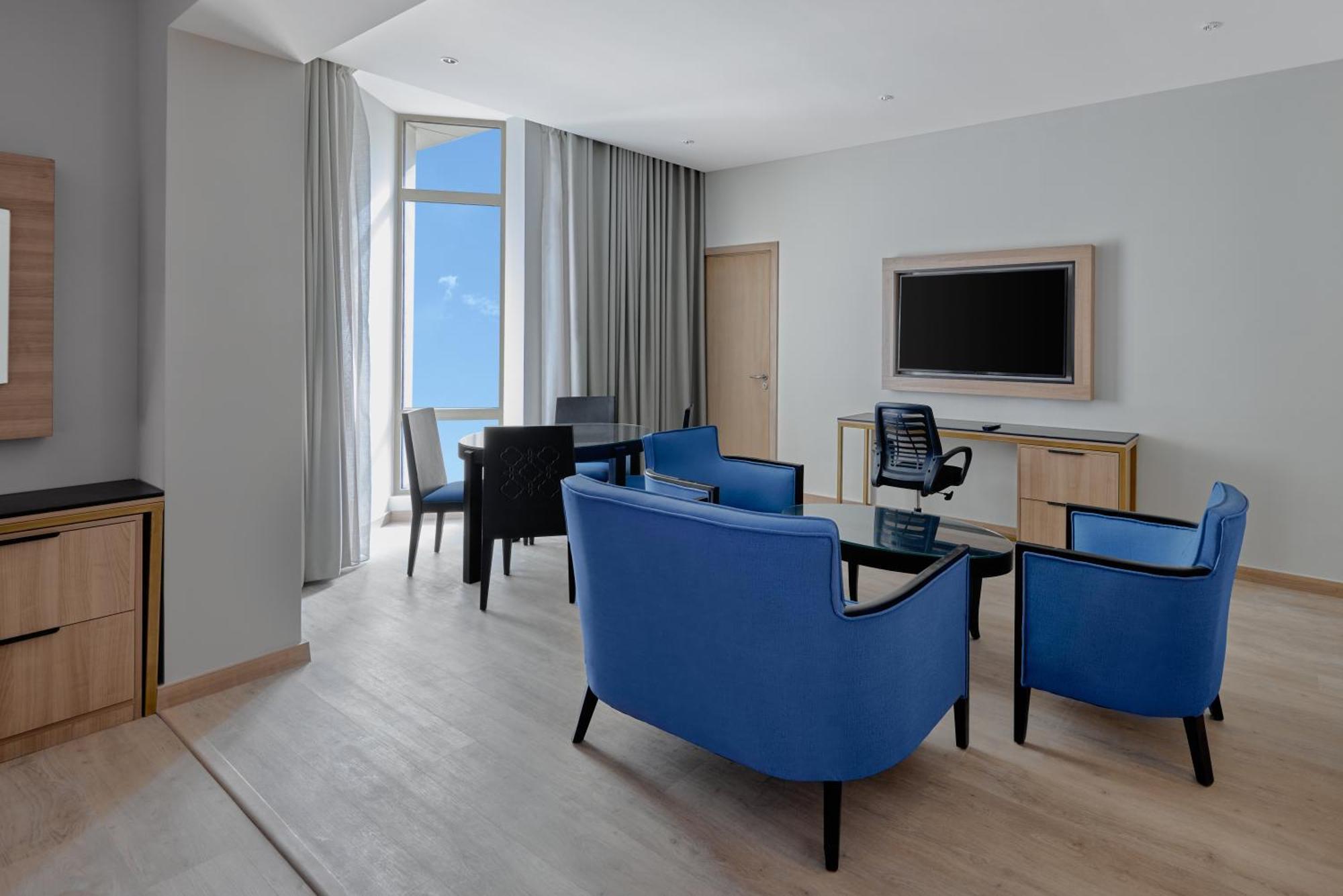 Makarem Umm Al Qura Hotel Mecca Exterior foto The photo shows a modern living area or hotel suite. It features a light-colored wooden floor and large windows allowing natural light to flood in. There are blue upholstered chairs arranged around a glass coffee table. A black office chair is positi