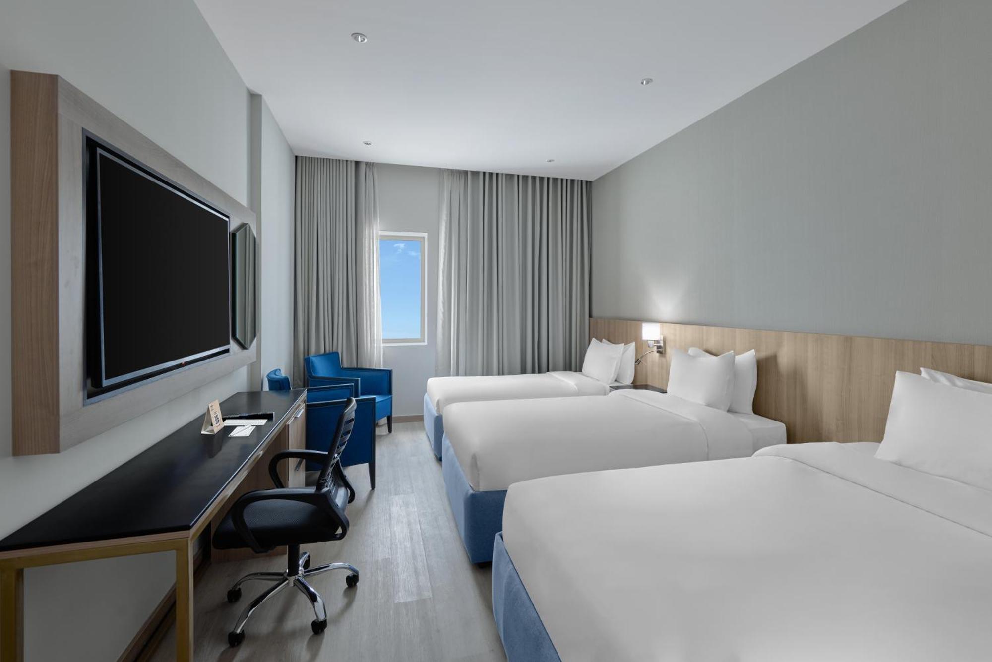 Makarem Umm Al Qura Hotel Mecca Exterior foto The photo shows a modern hotel room featuring two twin beds with white bedding. There's a light wood paneling behind the beds, and a large flat-screen TV mounted on the wall across from the beds. A comfortable blue armchair and a small desk with an e