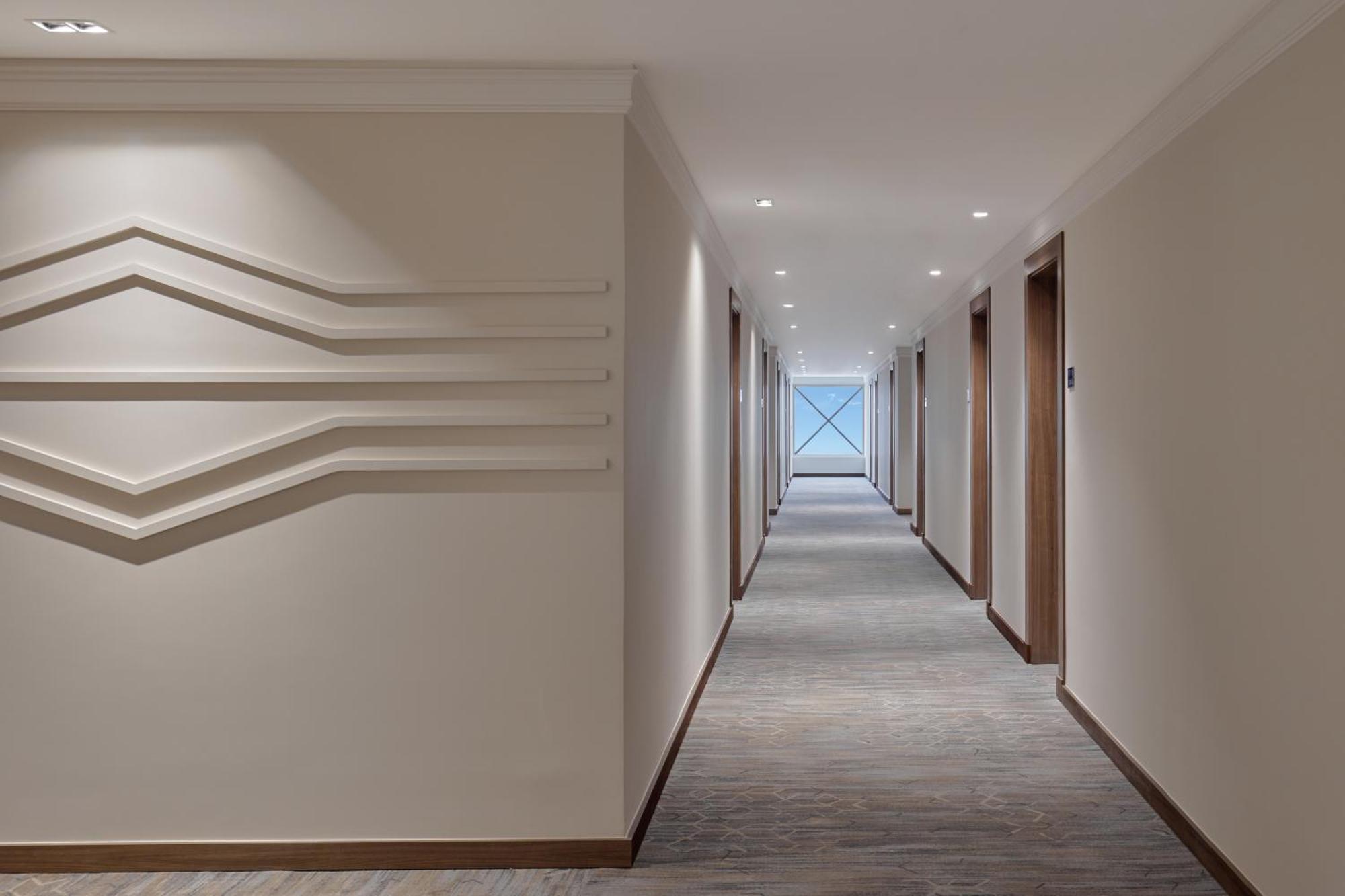 Makarem Umm Al Qura Hotel Mecca Exterior foto The photo shows a modern hotel corridor with a neutral color scheme. The walls are light-colored, and there are sleek, minimalistic features, including decorative lines on the left wall. The corridor itself is well-lit with recessed ceiling lights an