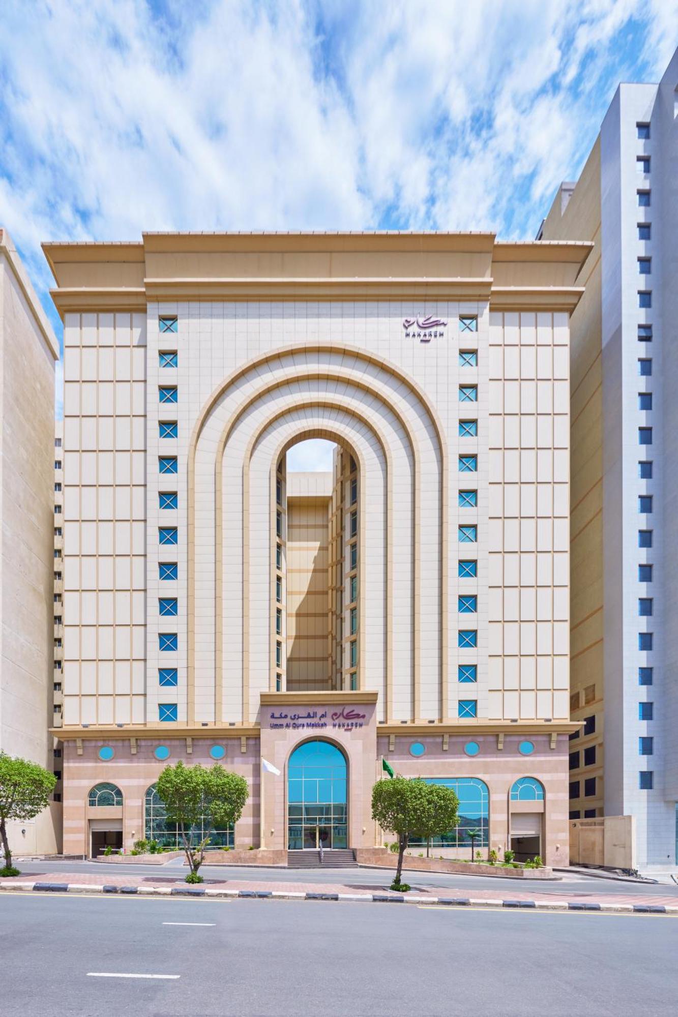 Makarem Umm Al Qura Hotel Mecca Exterior foto The photo shows a tall, modern building with a distinctive architectural design. The facade features a series of large arches and has a light-colored exterior with decorative patterns. There are blue accents that provide contrast against the neutral 