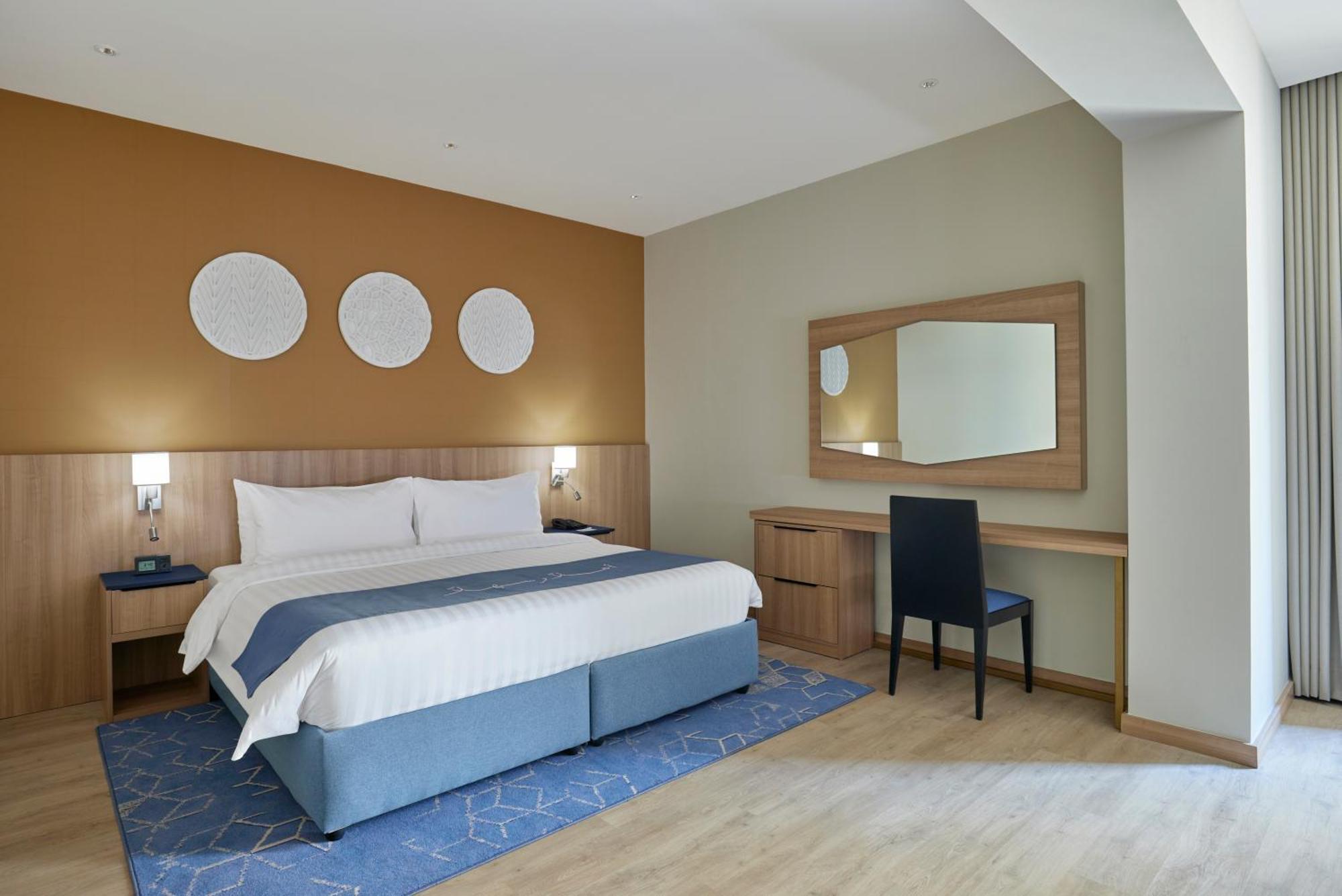Makarem Umm Al Qura Hotel Mecca Exterior foto The photo shows a modern hotel room. It features a large bed with white linens and a blue base. On the wall above the bed are three circular decorative pieces. There is a desk with a mirror next to it, and a black chair is positioned in front of the 