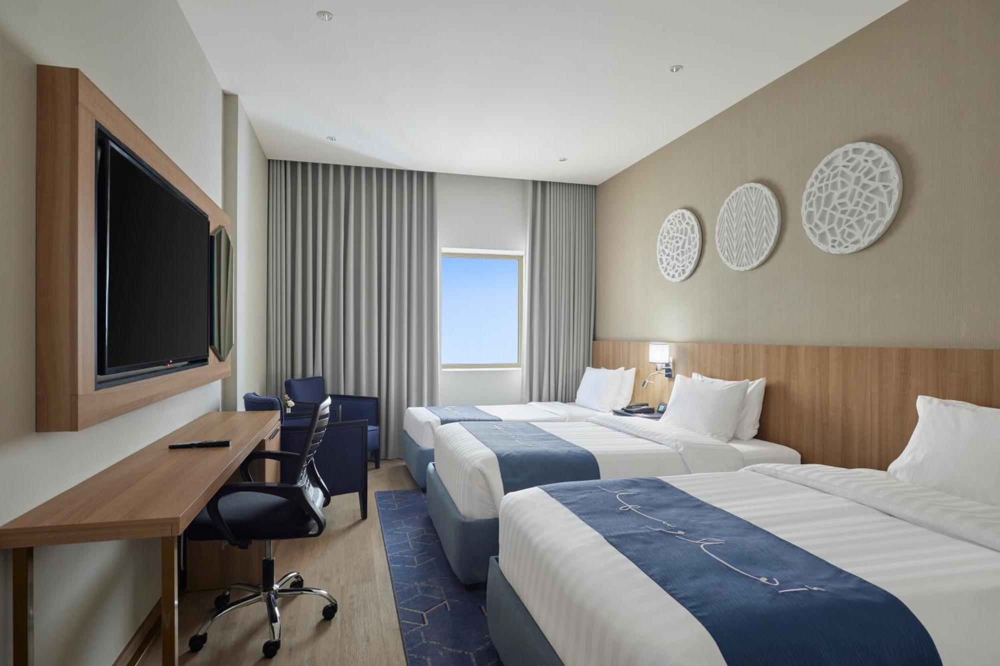 Makarem Umm Al Qura Hotel Mecca Exterior foto The photo shows a modern hotel room with two beds, each made with white linens and blue accents. There is a wooden wall with decorative circular pieces above the beds. A large television is mounted on the wall across from the beds. On the right side,