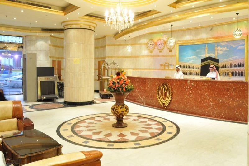 Makarem Umm Al Qura Hotel Mecca Exterior foto The photo shows an elegant hotel lobby. There are two receptionists behind a large, ornate front desk that has a marble finish. The lobby features a decorative circular area on the floor with a geometric pattern. In the center, there is a large vase 