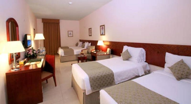 Makarem Umm Al Qura Hotel Mecca Exterior foto The photo shows a hotel room that features two sets of beds. The room is furnished with light-colored walls and a neutral color palette. Each bed has a light green and cream-colored bedspread. There’s a window with curtains, a desk with a chair, and 