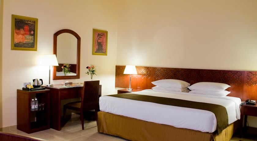 Makarem Umm Al Qura Hotel Mecca Exterior foto The photo shows a hotel room that features a double bed with a white and brown bedspread. There are bedside lamps on either side of the bed. Opposite the bed, there's a wooden desk accompanied by a chair, along with a mirror on the wall above the des