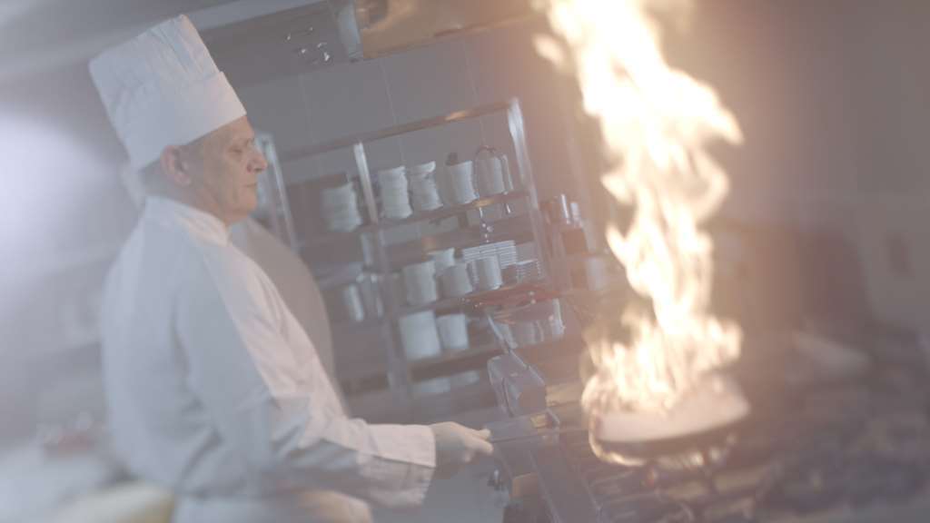 Makarem Umm Al Qura Hotel Mecca Restaurant foto The image shows a chef in a kitchen setting. He is wearing a white chef's uniform and a traditional chef's hat. There is a large flame erupting from a pan on the stove, which suggests that he is likely engaged in cooking that involves high heat or fl