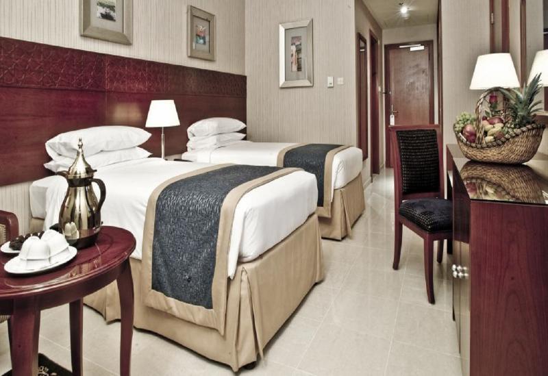 Makarem Umm Al Qura Hotel Mecca Exterior foto The photo shows a hotel room featuring two twin beds made with white linens and blue bed runners. A small bedside lamp is placed between the beds. On one side, there is a round table with a silver teapot and some small plates. To the right, there is 