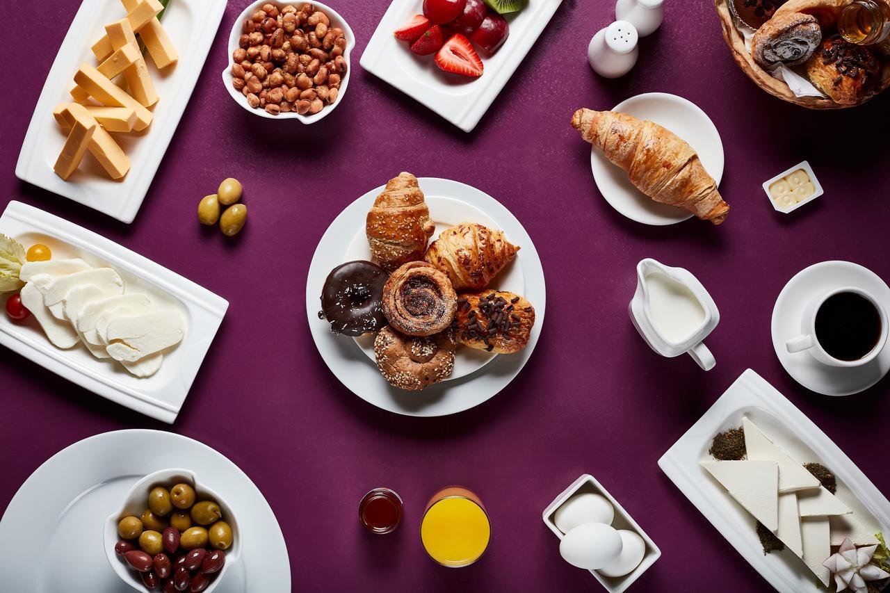 Makarem Umm Al Qura Hotel Mecca Exterior foto The photo displays a variety of breakfast foods arranged on a purple background. In the center, there's a plate with assorted pastries, including croissants and sweet rolls. Surrounding this plate are additional items: cubes of cheese, a bowl of oliv