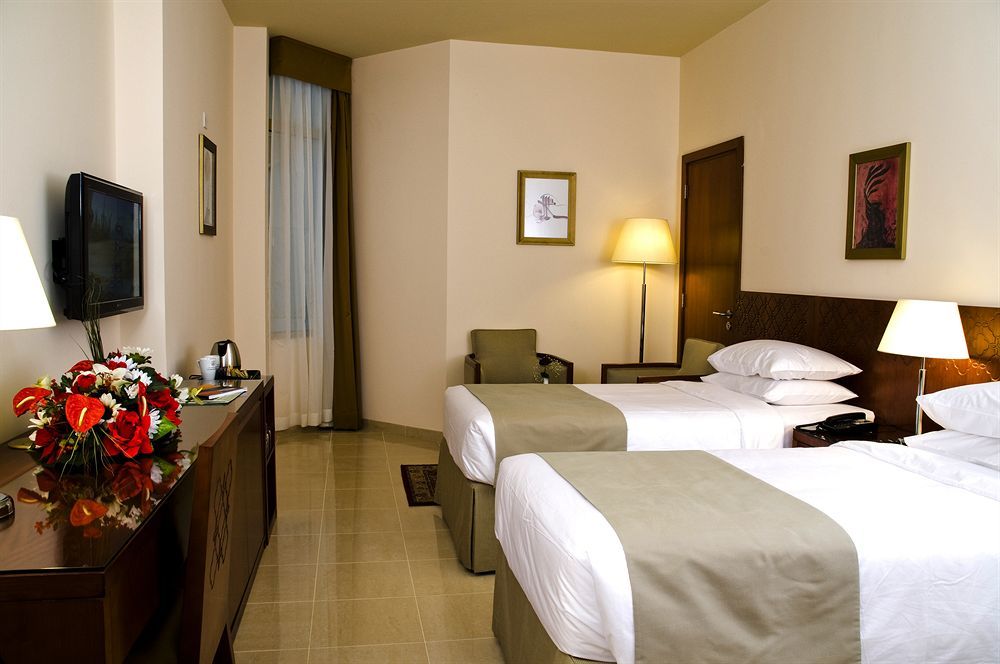 Makarem Umm Al Qura Hotel Mecca Exterior foto The photo shows a hotel room featuring two twin beds with white bedding and olive green accents. Next to the beds is a small bedside table with a lamp. There is a comfortable chair positioned nearby. A television is mounted on the wall, and there's a