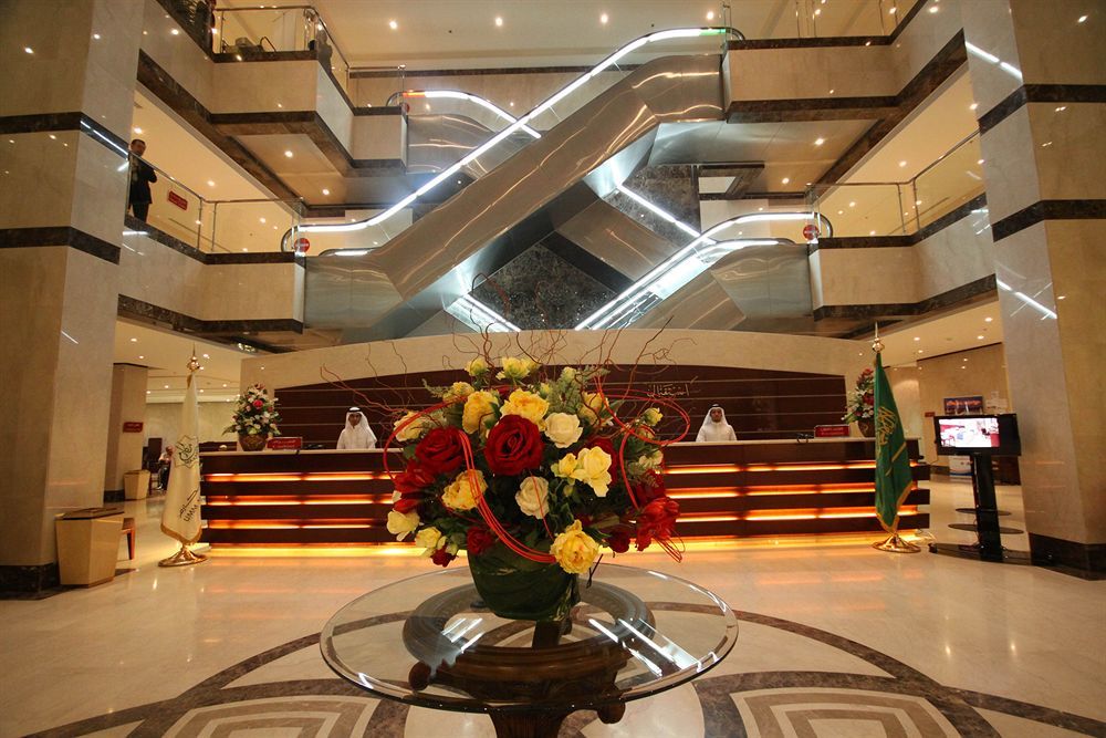 Makarem Umm Al Qura Hotel Mecca Exterior foto The photo shows a modern hotel lobby. In the foreground, there is an elegant floral arrangement featuring various colorful flowers in a decorative vase. In the background, a reception desk is visible, staffed by individuals in uniform. Two escalators