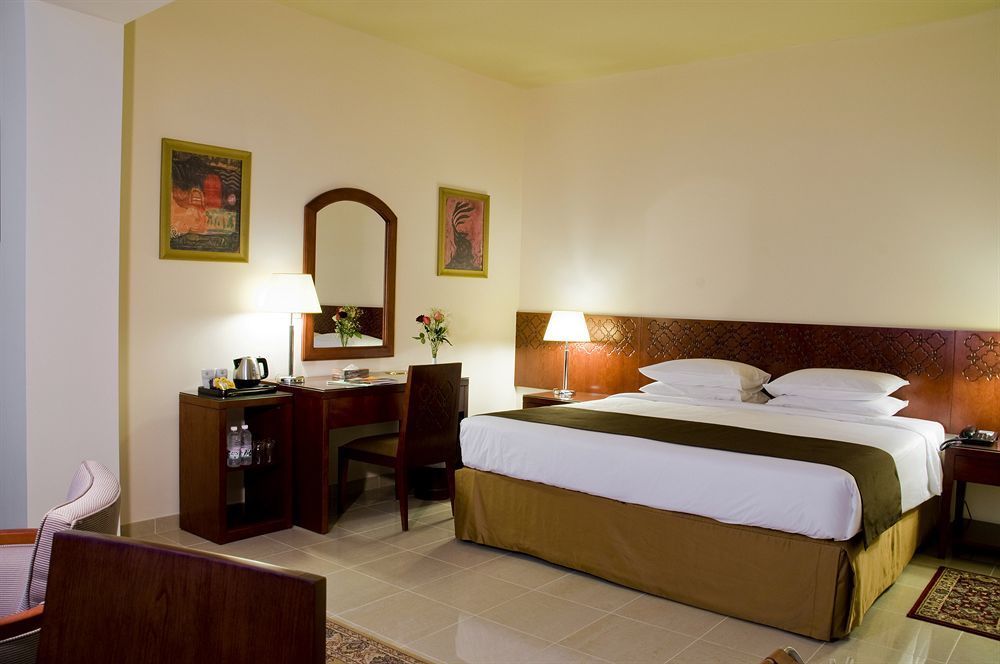 Makarem Umm Al Qura Hotel Mecca Exterior foto The photo shows a hotel room that features a neatly made bed with white linens and a brown bed skirt. On either side of the bed, there are bedside tables with lamps. A wooden desk and chair are present opposite the bed, accompanied by a small electro