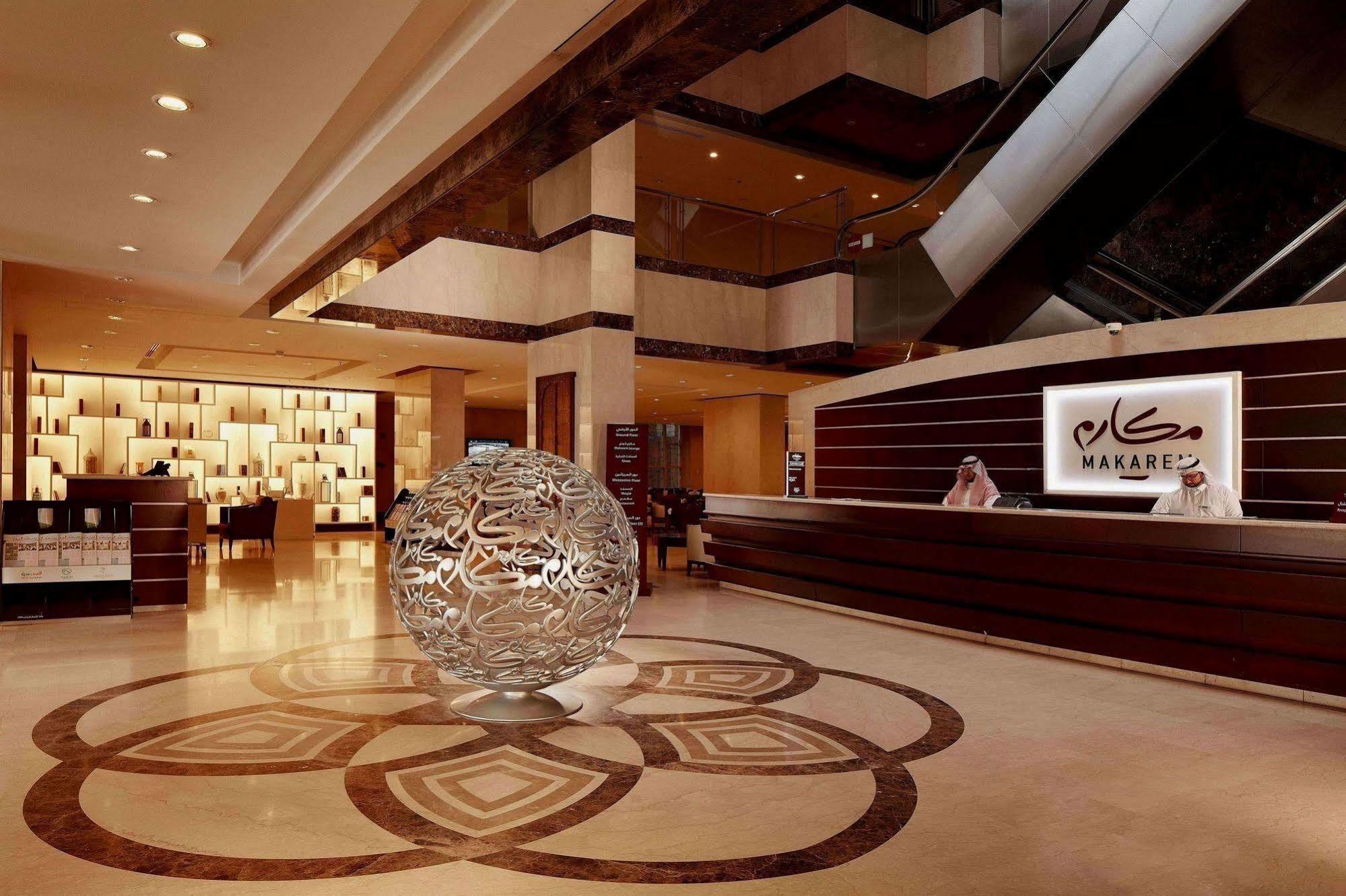 Makarem Umm Al Qura Hotel Mecca Exterior foto The photo shows an elegant hotel lobby featuring a modern and sophisticated design. In the center, there is a large decorative sphere with intricate patterns, placed on a stylized floor design. Behind the sphere, the reception desk is visible with st