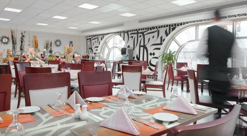 Makarem Umm Al Qura Hotel Mecca Exterior foto The photo shows the interior of a restaurant. It features a modern design with a combination of wooden and white furniture. The tables are set with white plates and folded napkins, arranged neatly. There are decorative touches such as small plants an