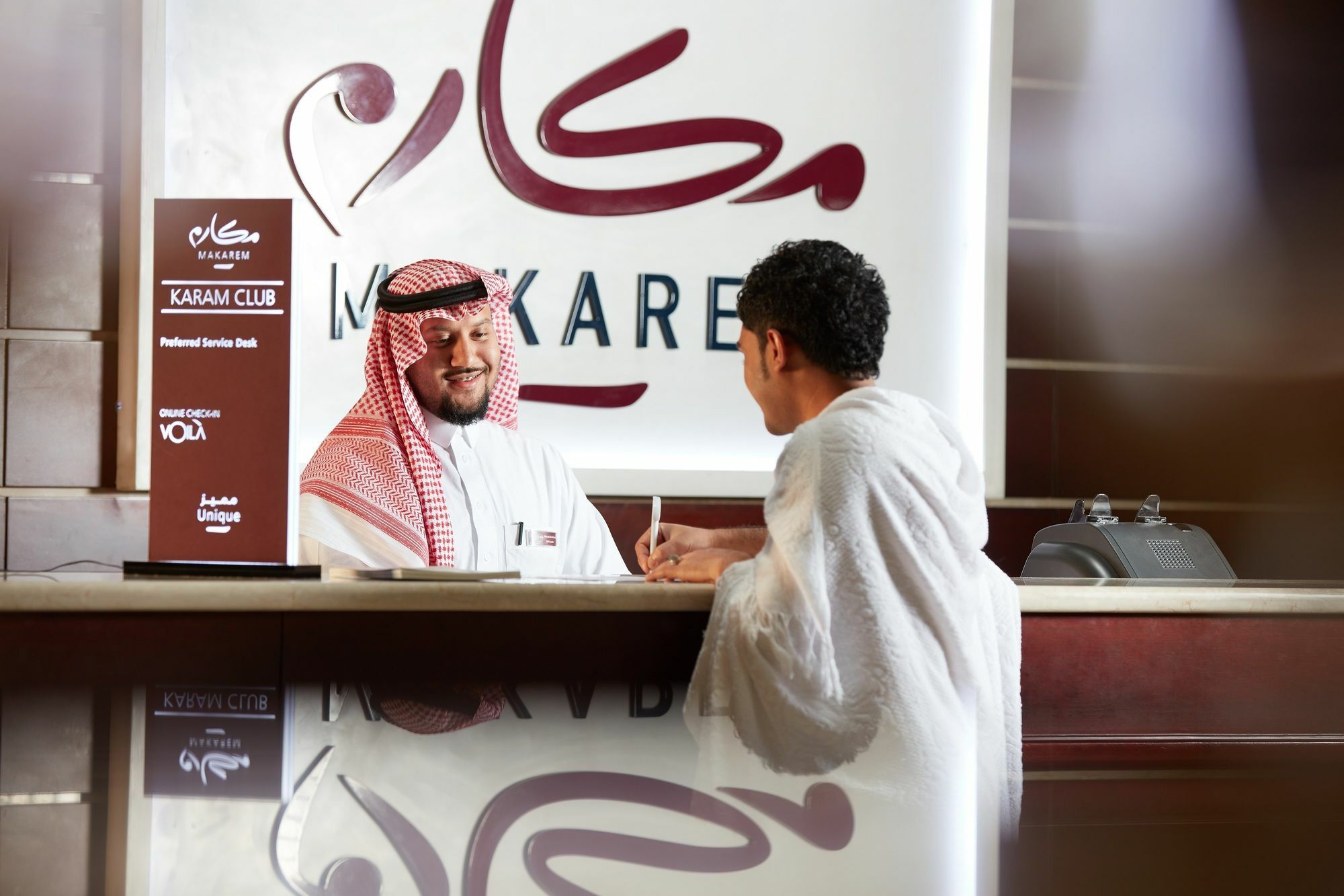 Makarem Umm Al Qura Hotel Mecca Exterior foto The photo shows a scene at a reception area. There are two men, one dressed in traditional Saudi attire with a red and white keffiyeh, who appears to be a staff member at a service counter. The other man, wearing a white robe, is engaged in conversat