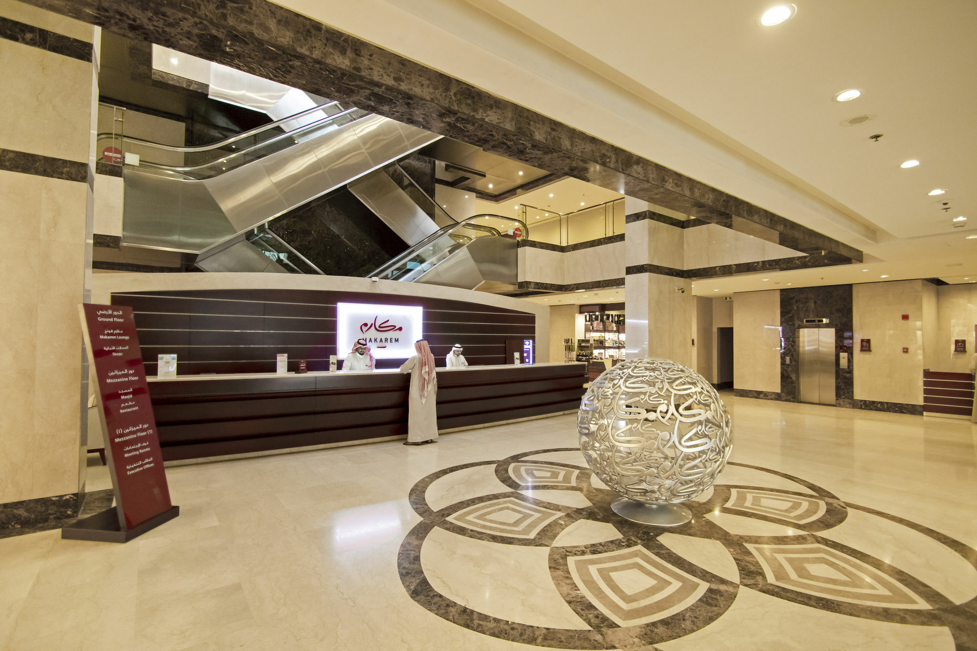 Makarem Umm Al Qura Hotel Mecca Exterior foto The photo shows the interior of a hotel lobby. There is a reception desk with staff attending to guests, and a modern, elegant design is evident throughout the space. In the center, there is a distinctive decorative spherical piece, likely made of gl