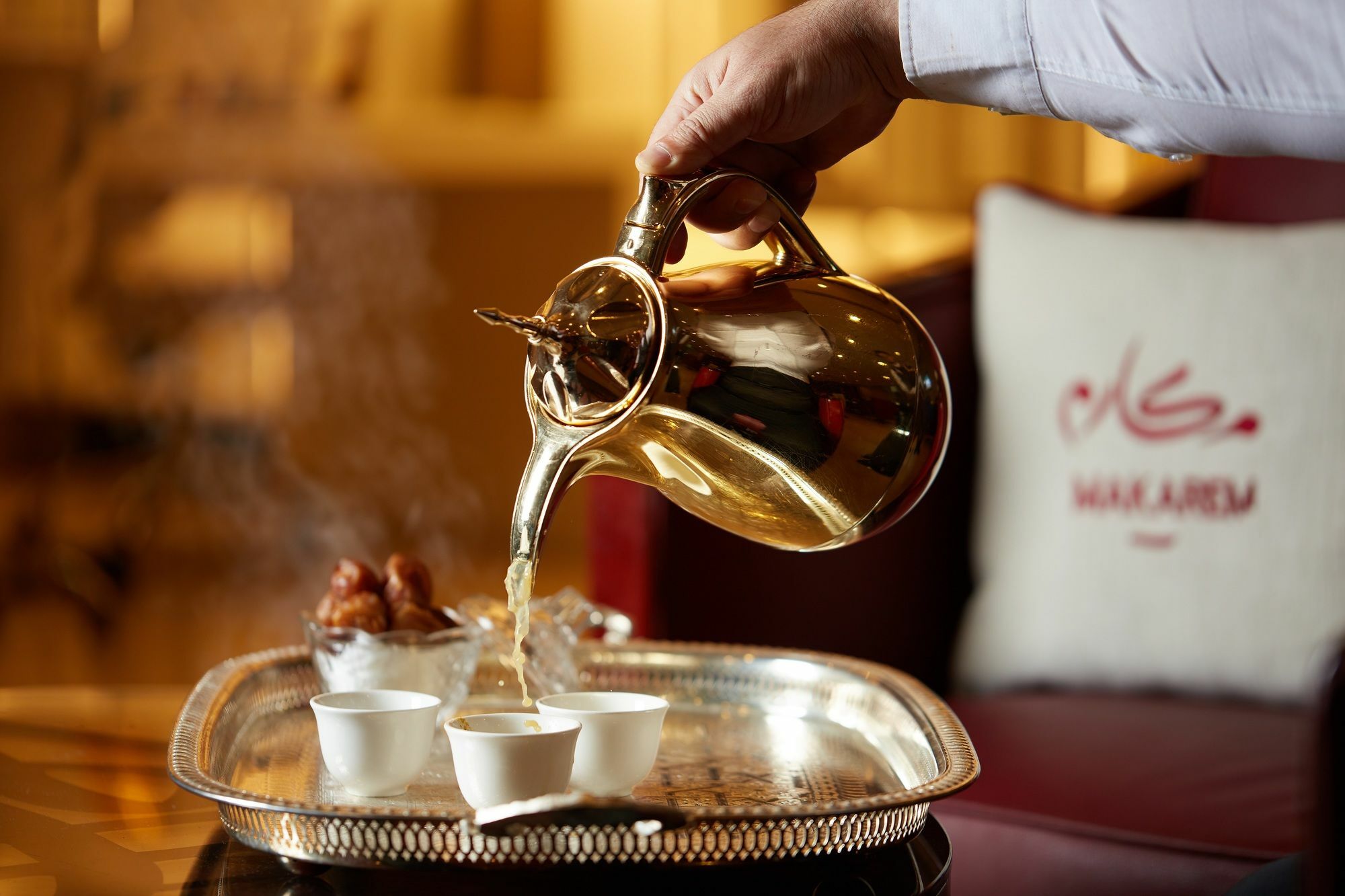Makarem Umm Al Qura Hotel Mecca Exterior foto The photo shows a hand pouring tea from a decorative pot into small white cups. The serving is taking place on a silver tray, which also includes dates. In the background, there is a cushion with Arabic script, suggesting a luxurious or traditional s